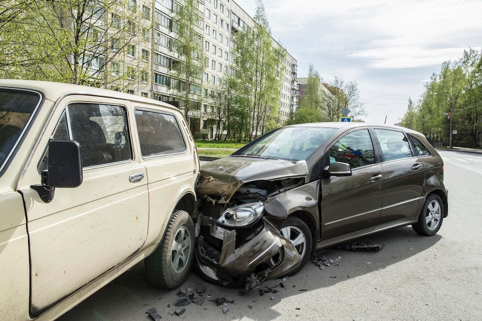 Two cars involved in a collision on a city street, with a damaged SUV and a sedan, surrounded by greenery and residential buildings, illustrating the risks of driving in urban areas.