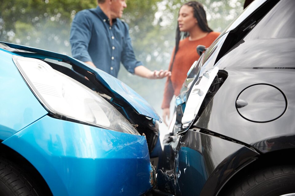 Two angry motorists arguing over responsibility after a car accident, with a damaged blue car and a black car in the foreground, set in a traffic-heavy environment.