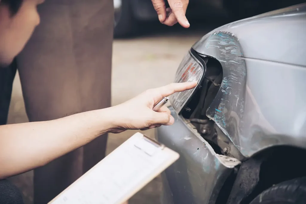 Person inspecting damage on a silver car, pointing at the rear light while holding a clipboard, related to car accident assessment and legal claims in Denton County.