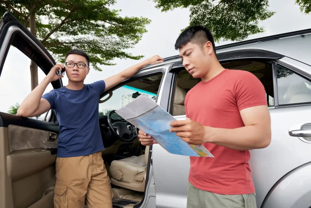 Two men discussing directions near a parked car, one on the phone and the other holding a map, in a setting suggesting a road trip or navigation challenge.