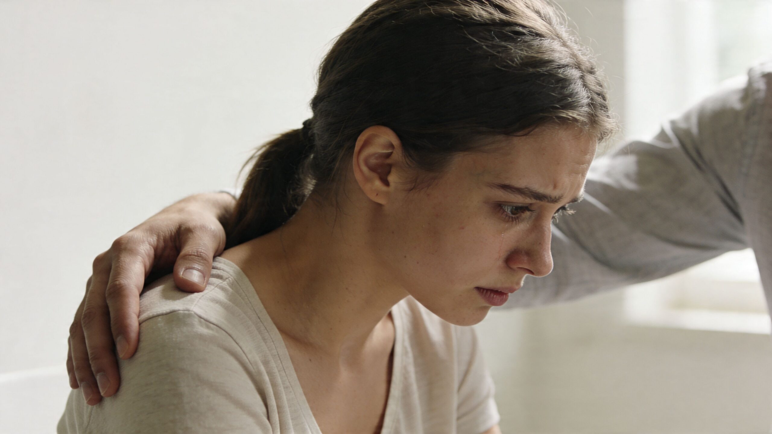 A distressed young woman receives comfort from a person placing a reassuring hand on her shoulder.